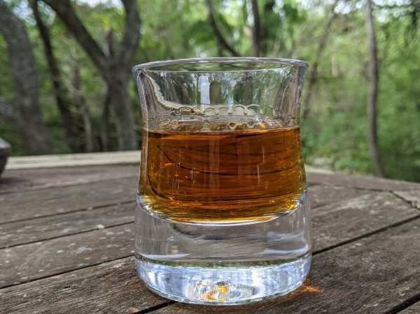 Amber Brown Rye Whiskey in a classic Rocks glass, on a picnic table with trees behind.