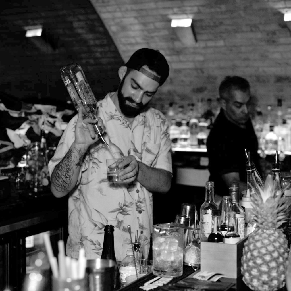 Danny Barragan. Black and White photograph of a bearded man mixing cocktails behind a bar wearing a Hawai'in shirt and ballcap.  Tools of the trade and a pineapple are in the foreground.
