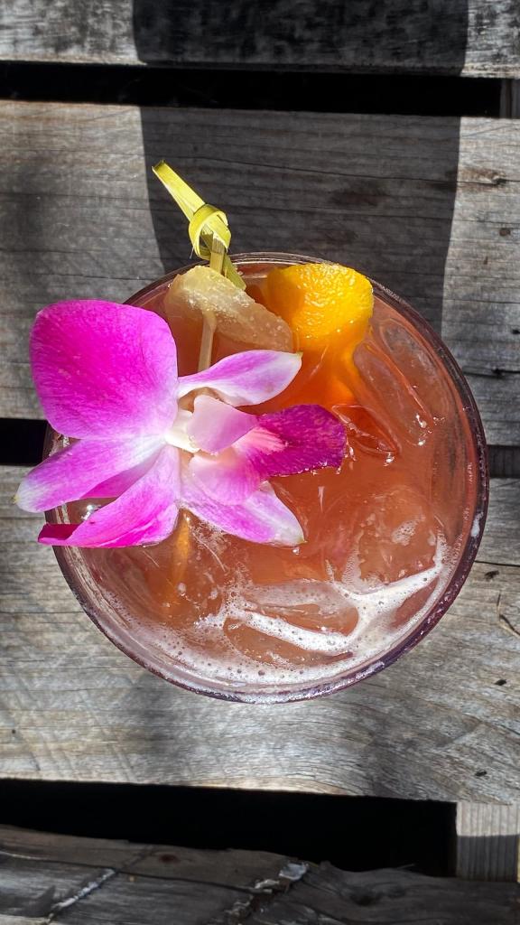 Wha'Sfumato You Tiki Cocktail, seen from directly above, setting on an outdoor table at the Tonga Hut in North Hollywood, CA. Cocktail is in a Rocks glass; blood-orange in color. It is garnished with a fuchsia and white orchid; a peel of orange zest, and a slice of candied ginger on a bamboo spear