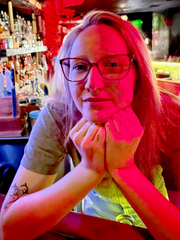 Photo of Emily Robin, Tiki Bartender at Tonga Hut. Emily is photographed behind the bar at Tonga Hut, in a subtle and exotic red to orange glow. She rests her chin on her gently closed fists, looking directly at the camera, smiling.